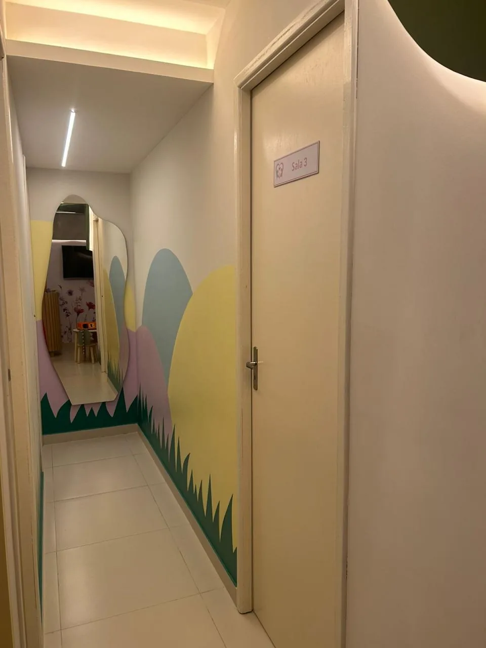 Corredor da clínica com parede pintada em morros coloridos, grama ilustrada e espelho orgânico