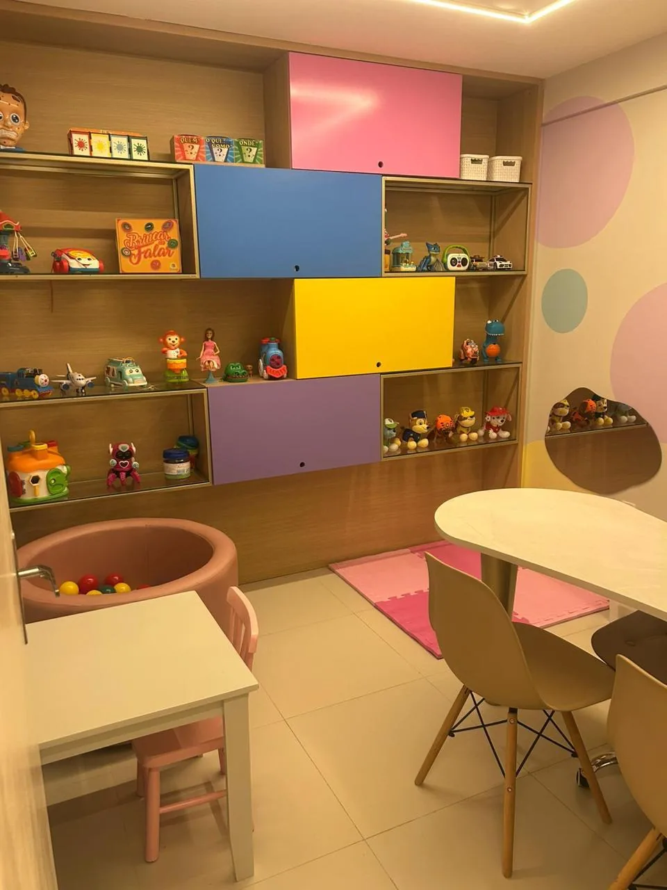 Sala de atendimento com estante de brinquedos e jogos de linguagem, piscina de bolinhas e mesa para família e terapeuta